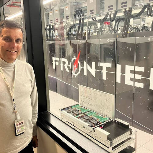 A smiling man stands beside a large window or glass wall, looking into the Oak Ridge National Laboratory data center where the “Frontier” supercomputer is installed behind panels labeled “FRONTIER.”